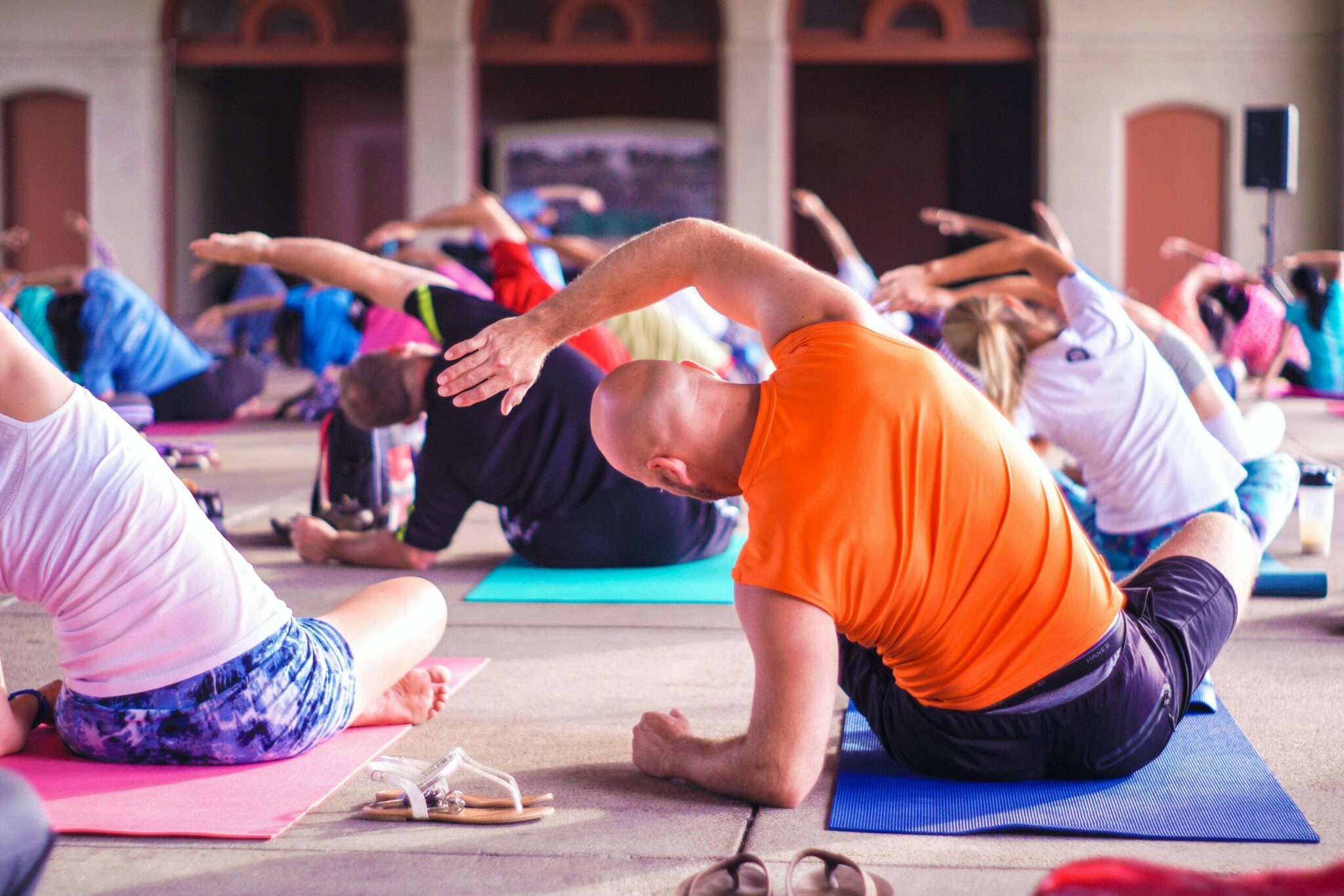 People in a yoga class are sitting on mats, leaning sideways with one arm over their heads. The scene is indoors, with a focus on movement and stretching—an ideal setting for tips for healthy living. Various colorful outfits add vibrancy to the room.