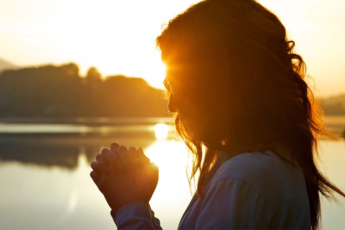 A person stands by a serene lake at sunset, hands clasped together in front, as the sun creates a warm glow around their silhouette. The peaceful scene captures a moment of reflection or prayer, perhaps contemplating their journey to overcome tramadol addiction.