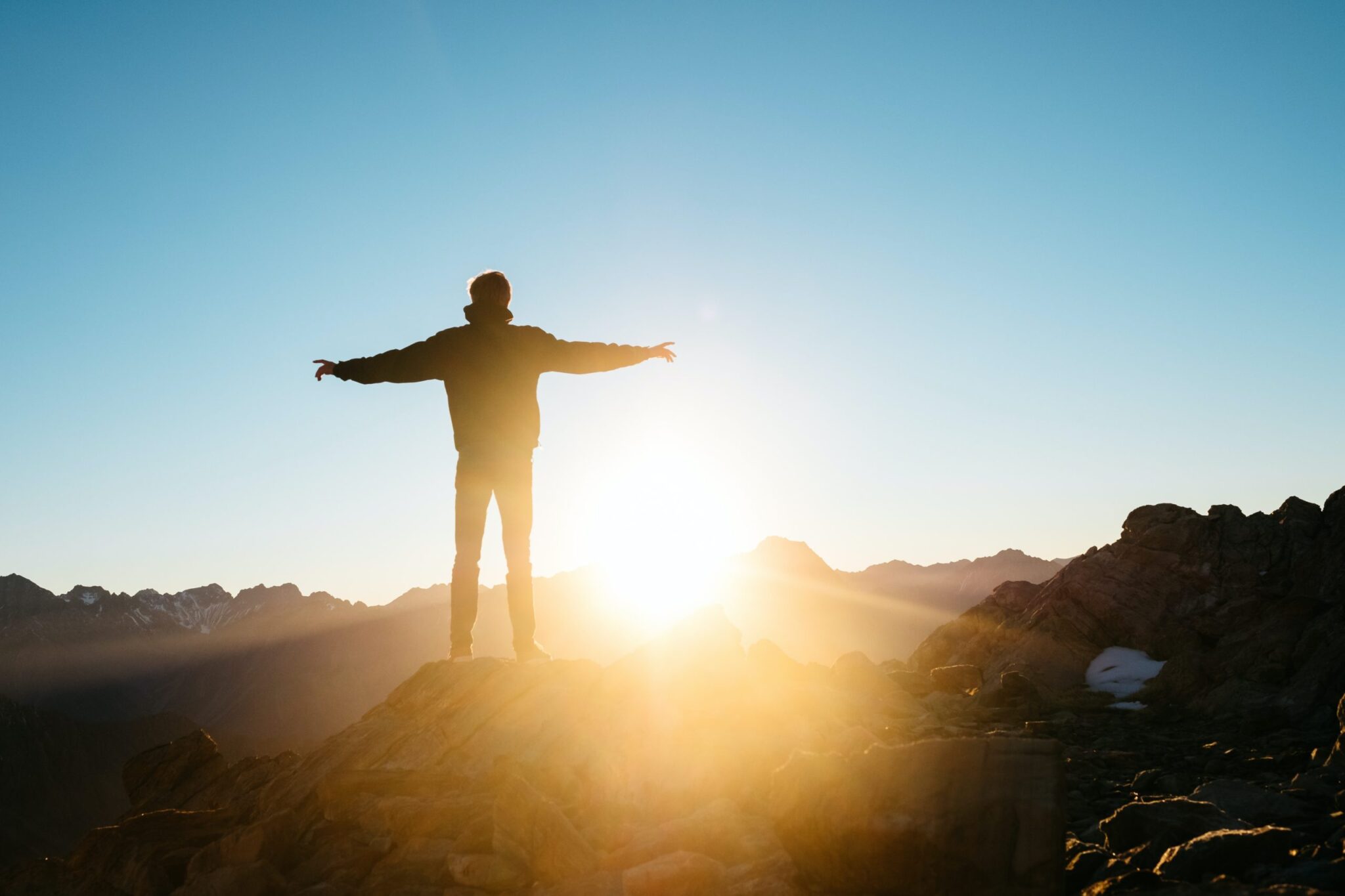 A person stands on a rocky peak with arms outstretched, silhouetted against a bright sun rising over distant mountain ranges under a clear blue sky, finding freedom in natures embrace—a powerful substitute for everyday addictions.
