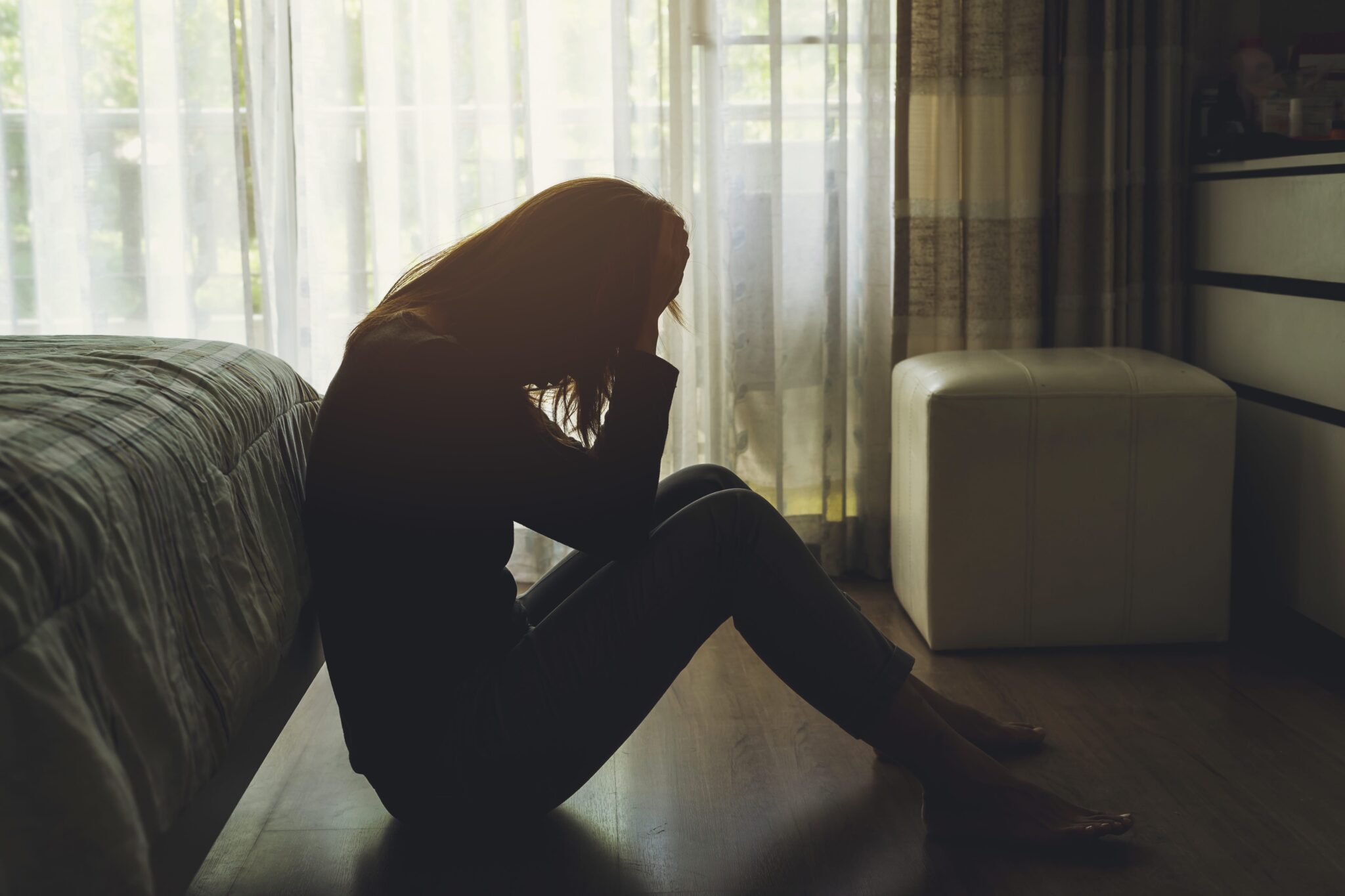 A person sits on the floor with knees drawn up, head in hands, silhouetted against a window with sheer curtains, capturing the somber mood often linked to cocaine use and depression.