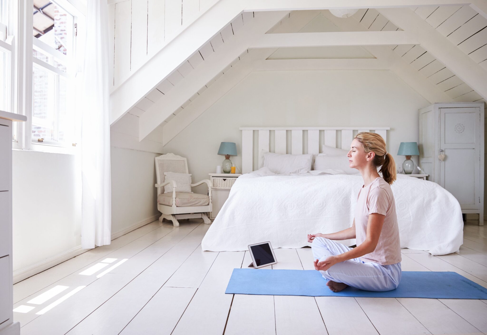 Woman sits cross-legged on a yoga mat meditating in a bright, white bedroom with a bed, chair, and tablet on the floor nearby—her peaceful routine to become a morning person.