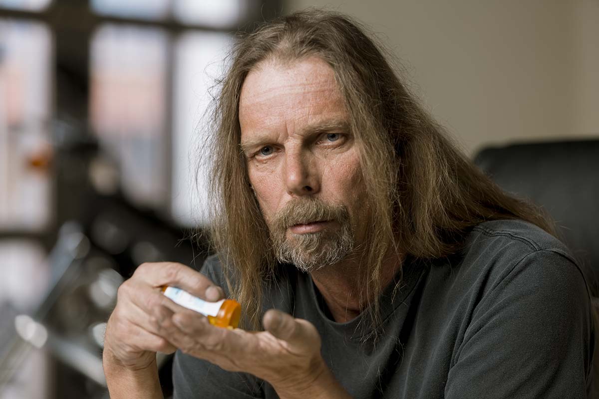 A middle-aged man with long hair and a beard sits indoors, looking serious while pouring pills from a prescription bottle into his hand—reflecting the reality behind troubling opioid addiction statistics.