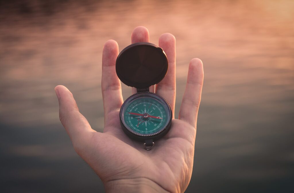 A hand holds an open compass against a blurred, warm-toned background. The compass needle points north, symbolizing direction and hope, much like finding ones path in addiction treatment. Evening light casts soft reflections on the water, creating a serene and contemplative atmosphere.