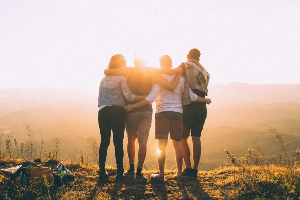 Four people stand arm in arm at the edge of a grassy hill, facing a sunset. They overlook a vast, serene landscape, finding solace as the sun casts a warm, golden light. In this tranquil moment, they gather strength to resist any relapse into past troubles.