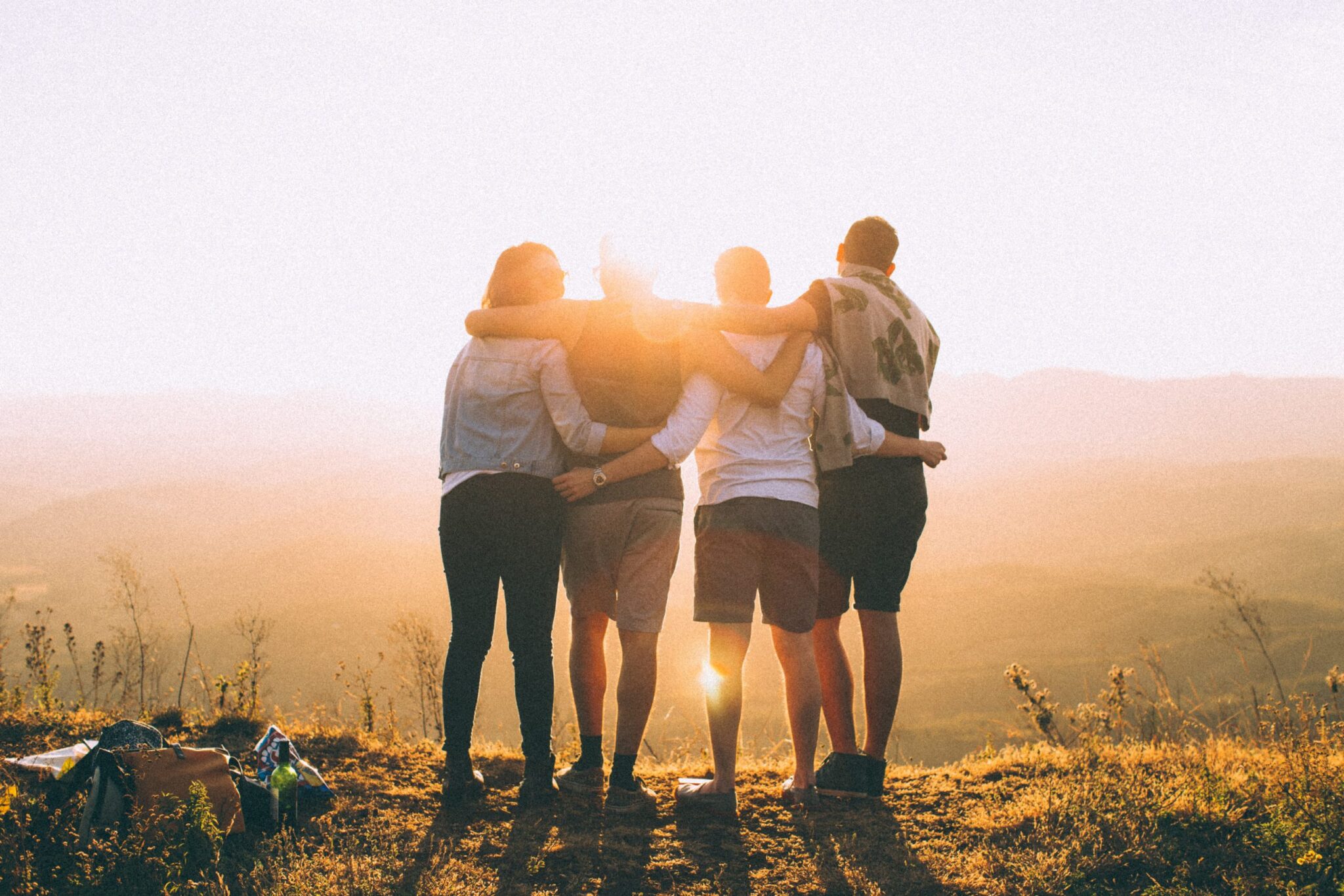 Four people stand arm in arm at the edge of a grassy hill, facing a sunset. They overlook a vast, serene landscape, finding solace as the sun casts a warm, golden light. In this tranquil moment, they gather strength to resist any relapse into past troubles.