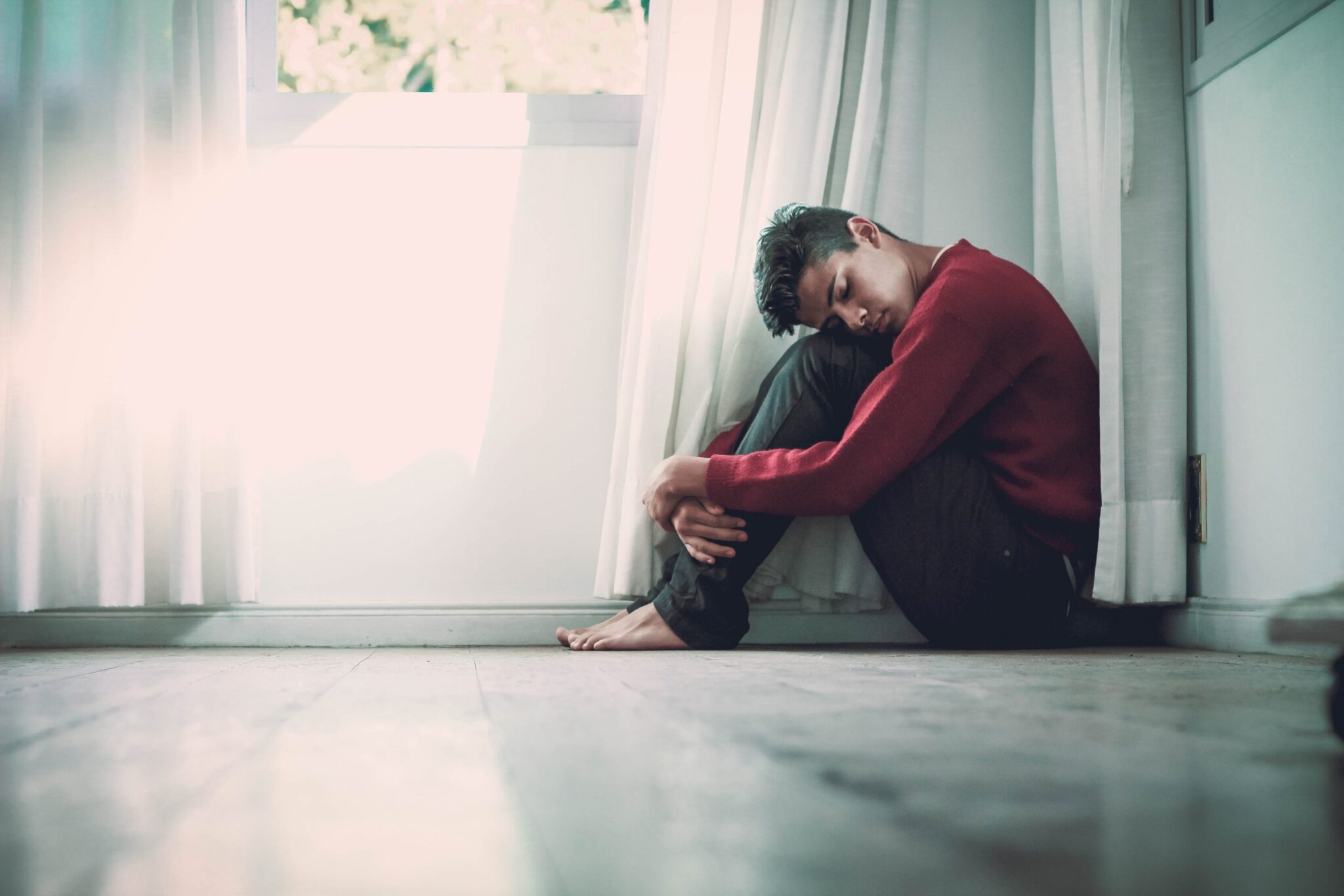 A person in a red sweater sits on a wooden floor, hugging their knees near a window with sheer white curtains. Sunlight filters through the curtains, casting a gentle glow in the room.