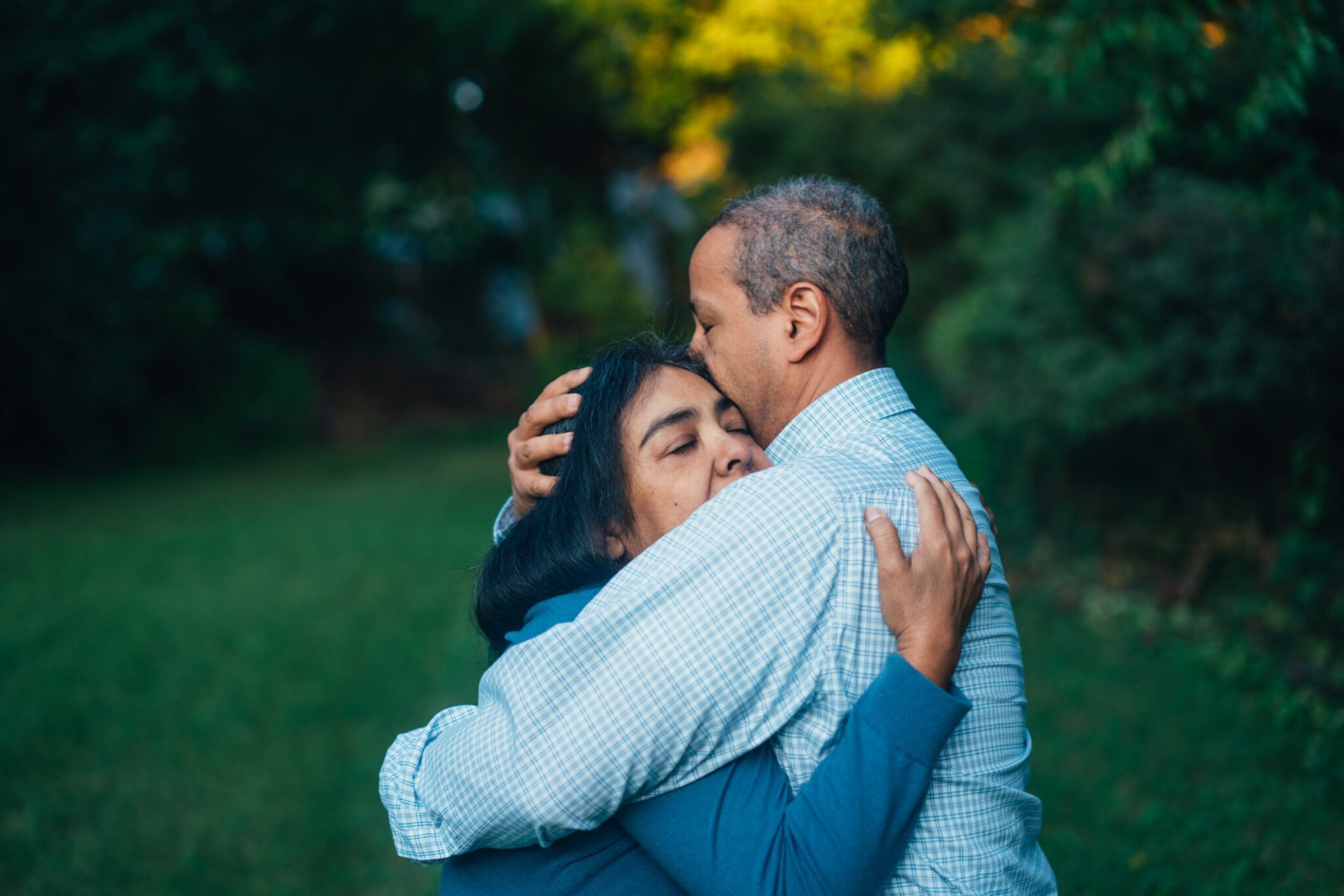 A man and woman embrace in a green park. The woman, wearing a blue top, has her eyes closed, resting her head on the mans chest. The man, in a checked shirt, holds her closely, with his eyes closed, surrounded by lush greenery.