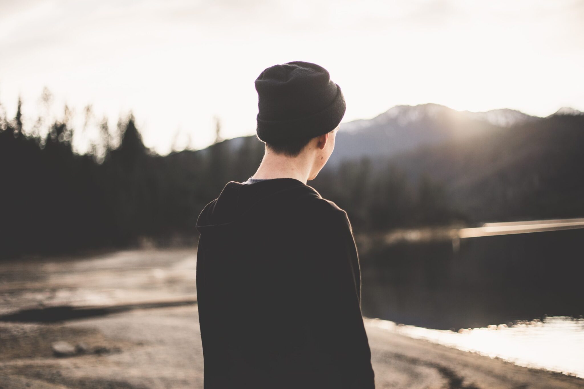 A person wearing a beanie and dark jacket is standing by a calm lake, contemplating the distant mountains under a clear sky. The serene landscape, free from any signs of alcoholism, includes a sandy shore and trees lining the waters edge.