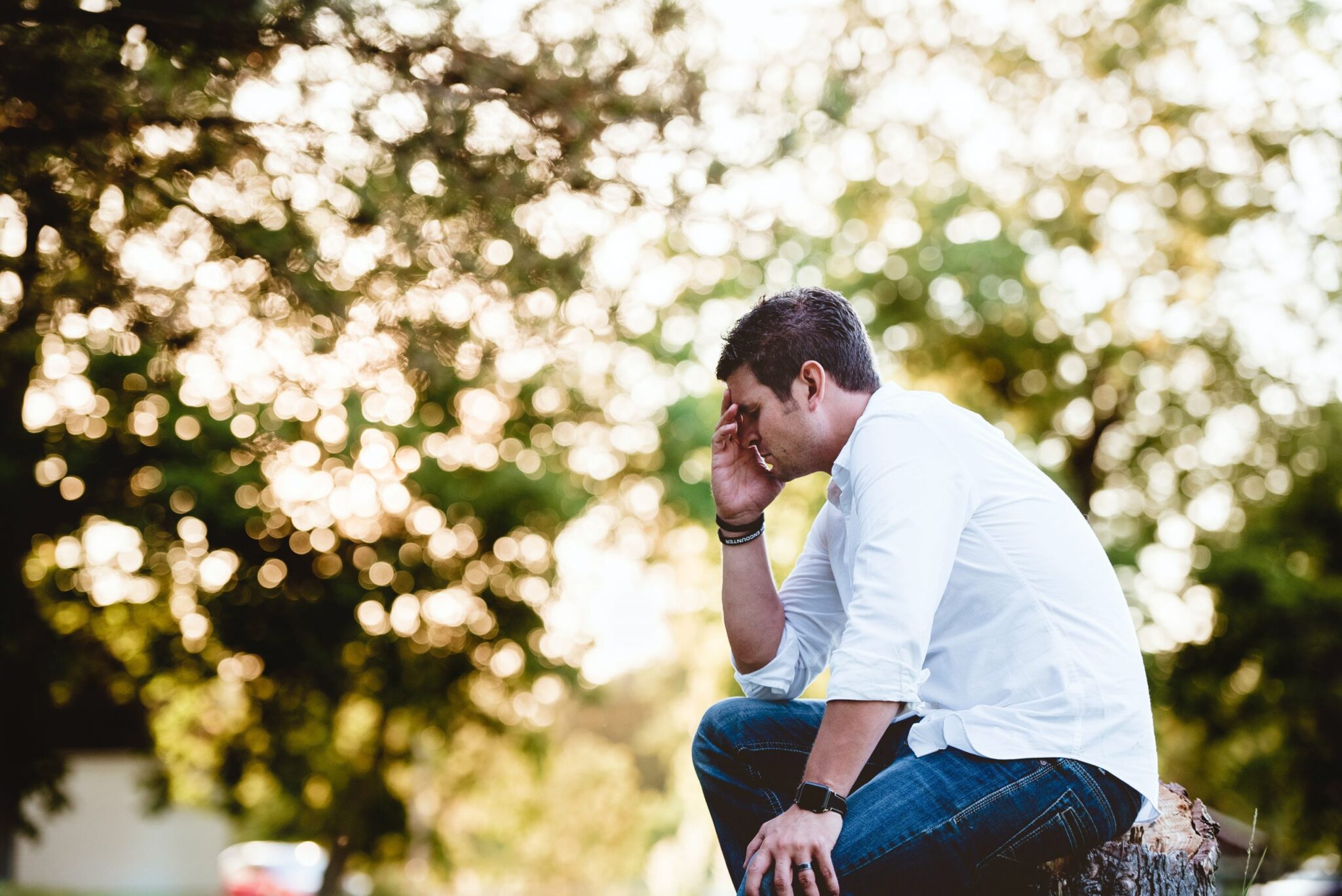 A person in a white shirt and jeans sits on a tree stump in a park, holding their forehead with one hand. The background is blurred with sunlight filtering through the trees, suggesting signs of alcoholism as they appear to be lost in deep thought or contemplation.