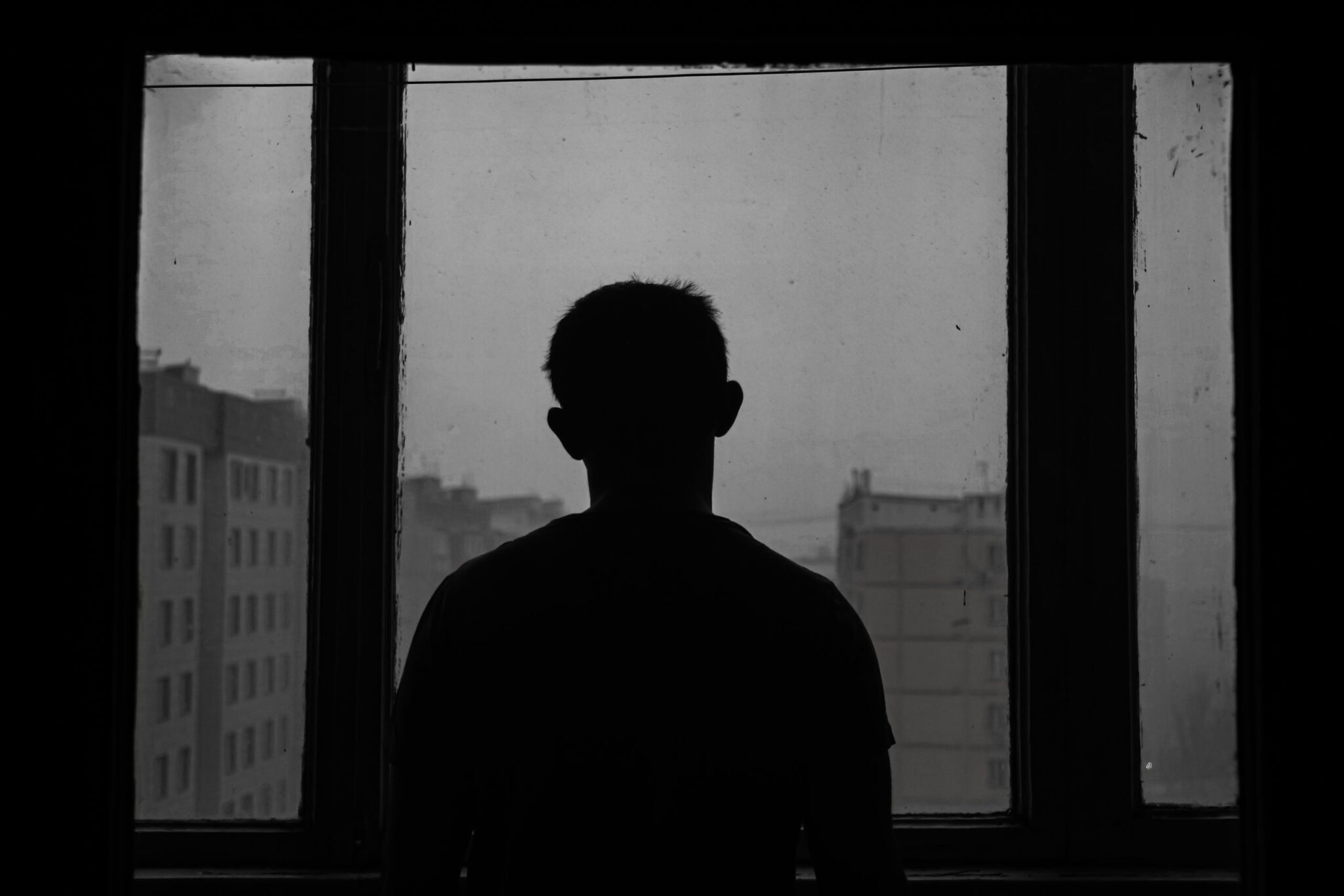 Silhouette of a person standing in front of a window, gazing outside at a row of buildings. The scene is in grayscale, evoking a somber and reflective mood, hinting at possible signs of alcoholism hidden beneath the surface.