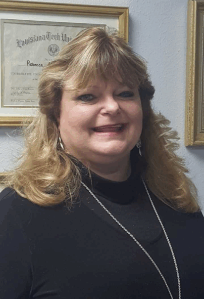 A woman with shoulder-length blonde hair, wearing a black top and a necklace, smiles at the camera. Behind her, a framed diploma from Louisiana Tech University hangs on a light-colored wall.
