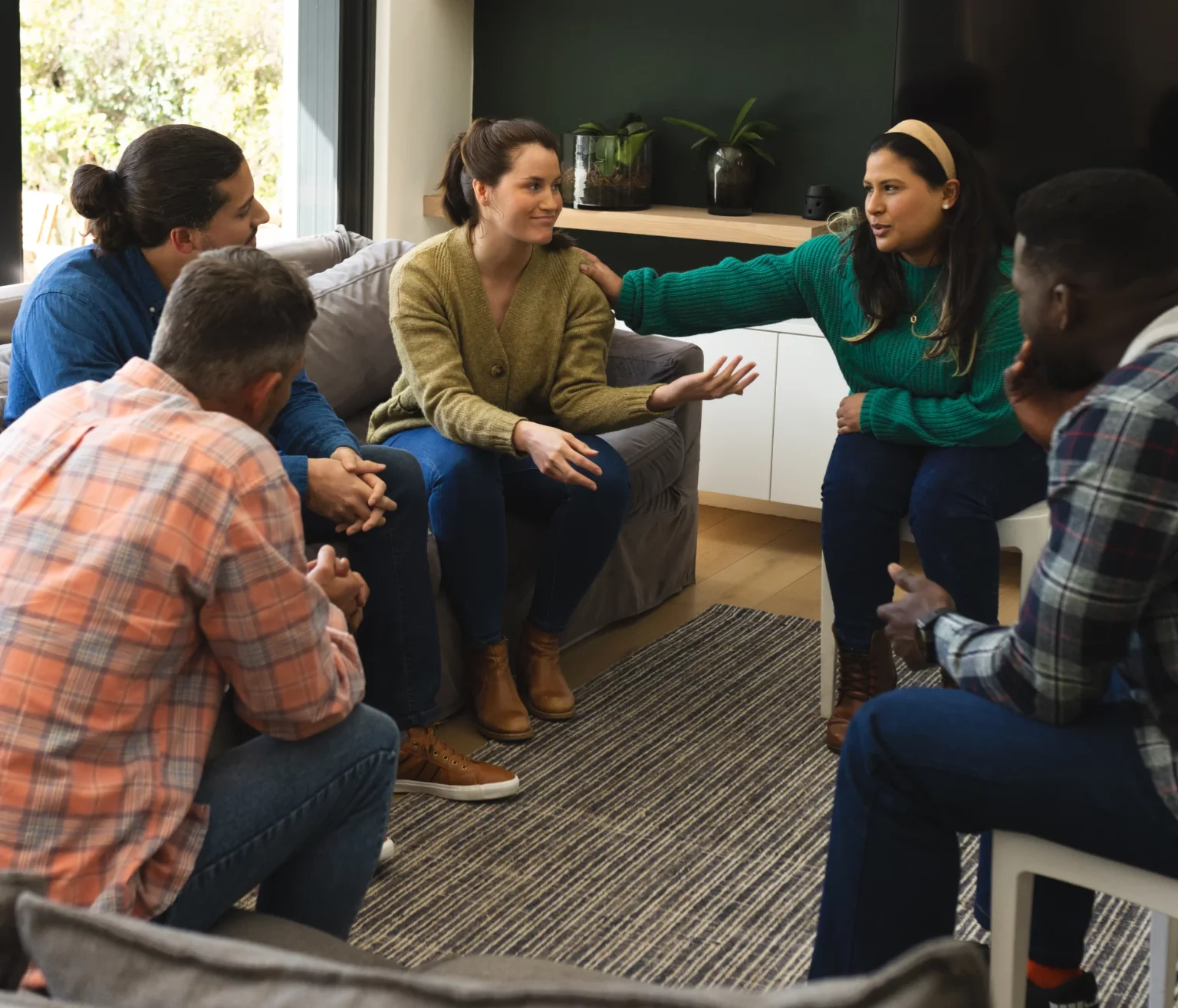 Five people sit in a living room having a serious group discussion; one woman gestures with her hand while another places a hand on her shoulder.