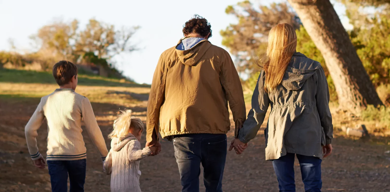 Four people, two adults and two children, are walking outdoors on a dirt path, holding hands, with their backs to the camera. Trees and grass are visible in the background.