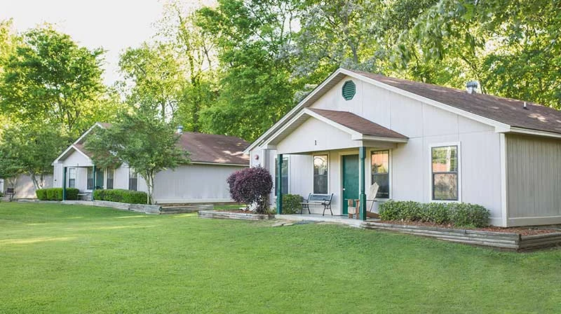 Single-story houses with front porches sit on a well-maintained lawn, surrounded by trees and landscaping, creating a peaceful setting ideal for a Treatment Center on a sunny day.