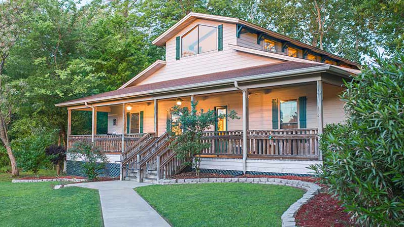 A two-story house with a large front porch, wooden railings, and green shutters, surrounded by trees and landscaped yard.