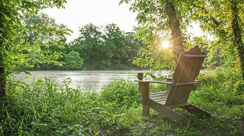 A wooden Adirondack chair faces a calm river, surrounded by green foliage, with sunlight shining through the trees in the background.