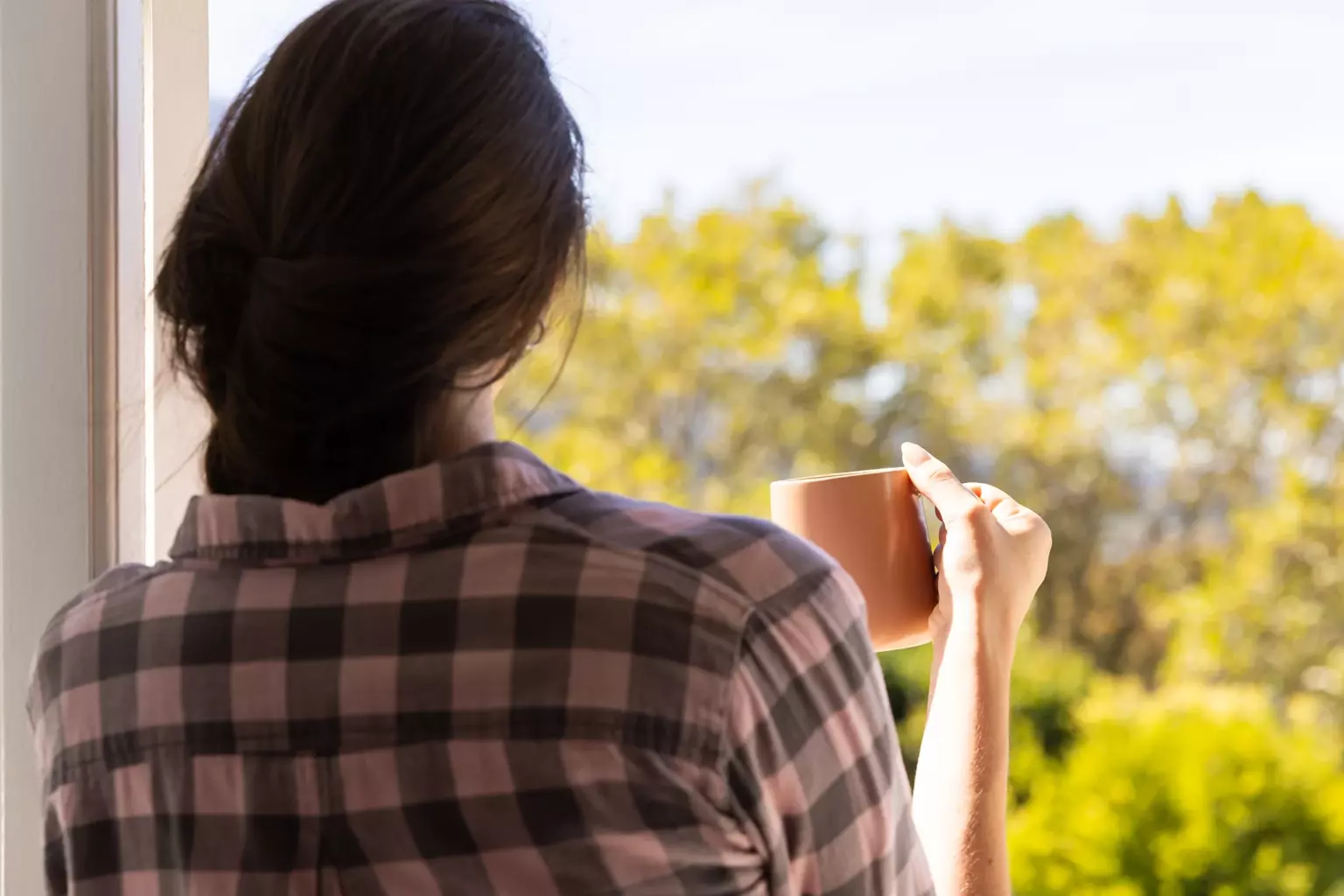 A person with long hair in a plaid shirt stands facing outdoors, holding a mug, and looking at trees and greenery in daylight.
