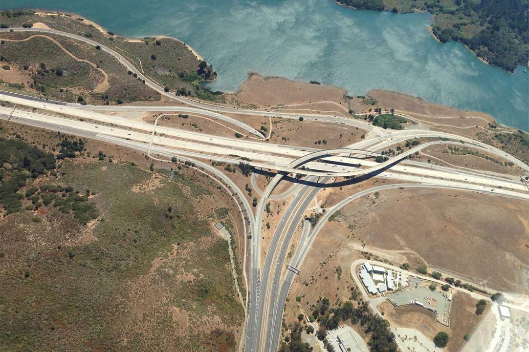 Aerial view of a freeway interchange with curved ramps and overpasses near a body of water and surrounding dry land.