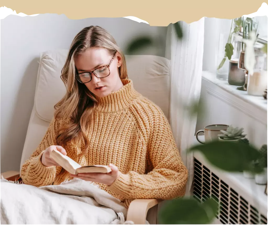 A woman wearing glasses and a chunky yellow sweater sits in a chair by a window, reading a book with a neutral expression.