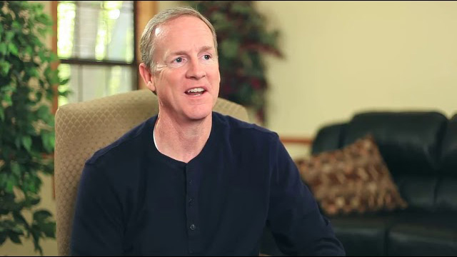 A middle-aged man in a dark shirt sits in a chair, smiling, with a sofa and plants visible in the background of a living room.