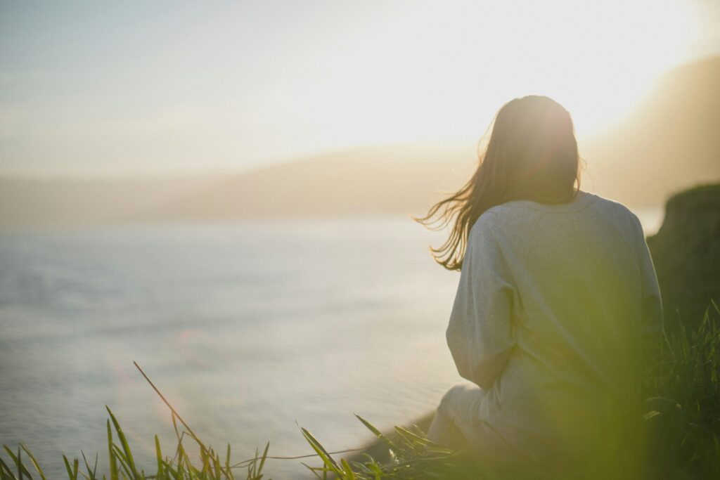 A person with long hair sits on grass facing a body of water, sunlight streaming in from behind, creating a peaceful and serene atmosphere—an ideal setting to reflect after an intensive outpatient program.