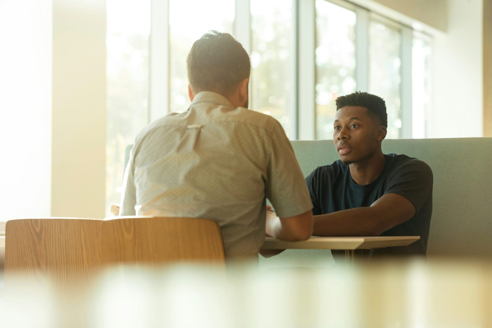 Two men are sitting across from each other at a table in a bright, modern setting, having a serious conversation about substance abuse treatment. One man faces the camera, listening attentively, while the other has his back to the camera.