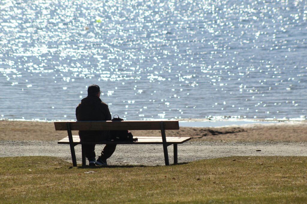 A person sits alone on a bench facing a sparkling body of water, sunlight reflecting off the surface. The calm, peaceful scene contrasts with their quiet struggle with depression and addiction, grass in the foreground and a sandy beach near the water’s edge.