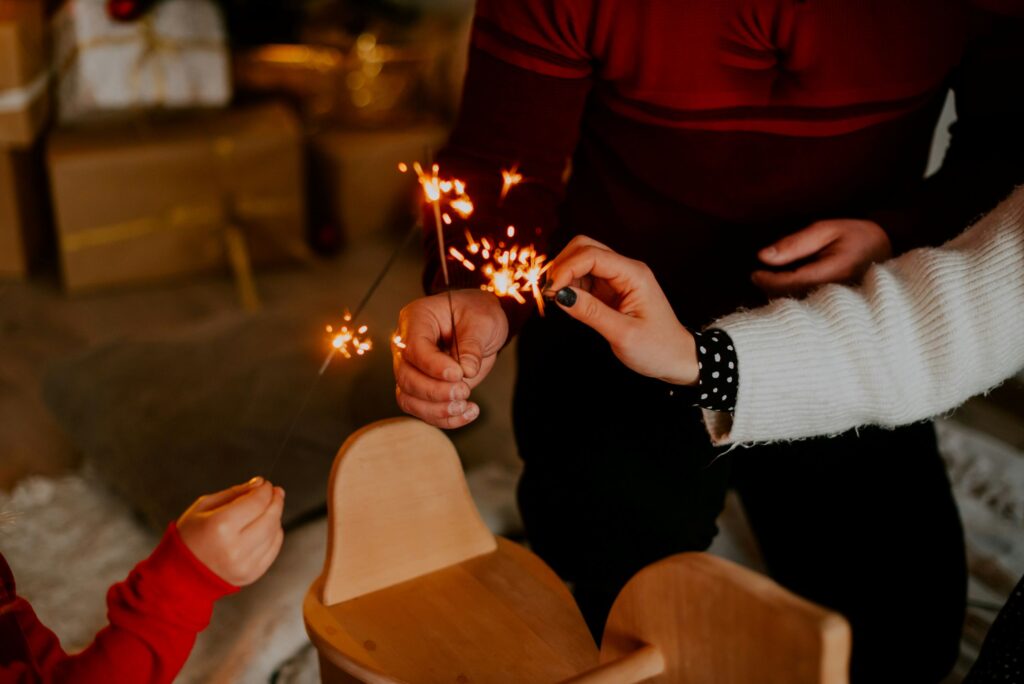 Three people holding sparklers together indoors near a wooden rocking horse, with presents and cozy blankets in the background, celebrate milestones in addiction recovery, creating a festive and warm atmosphere.