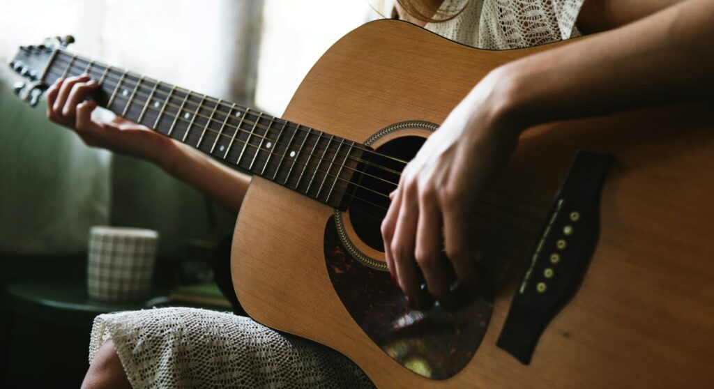 A person in early sobriety, wearing a light textured dress, plays an acoustic guitar indoors. One hand is on the fretboard and the other strums as a checkered mug sits nearby on a small table.