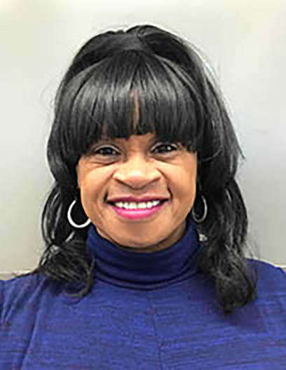 A woman with medium-length black hair and bangs, wearing hoop earrings and a blue turtleneck, smiles at the camera against a plain light background, displaying confidence as an MSW professional.