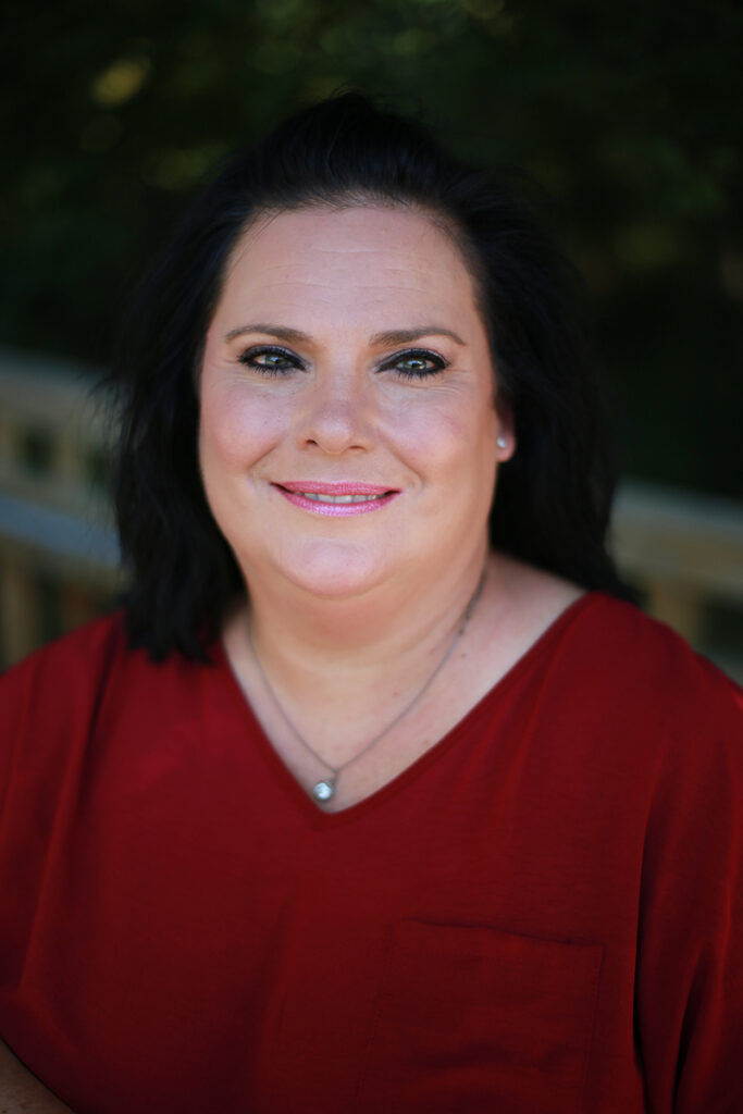 Belinda Kennedy, a woman with dark hair and a red top, smiles at the camera outdoors against a blurred green background.