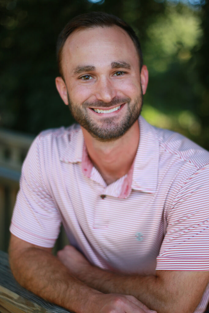 Brandon Waits, Ph.D., with short brown hair and a beard, smiles at the camera in a light pink striped polo shirt, seated outdoors amid lush greenery.