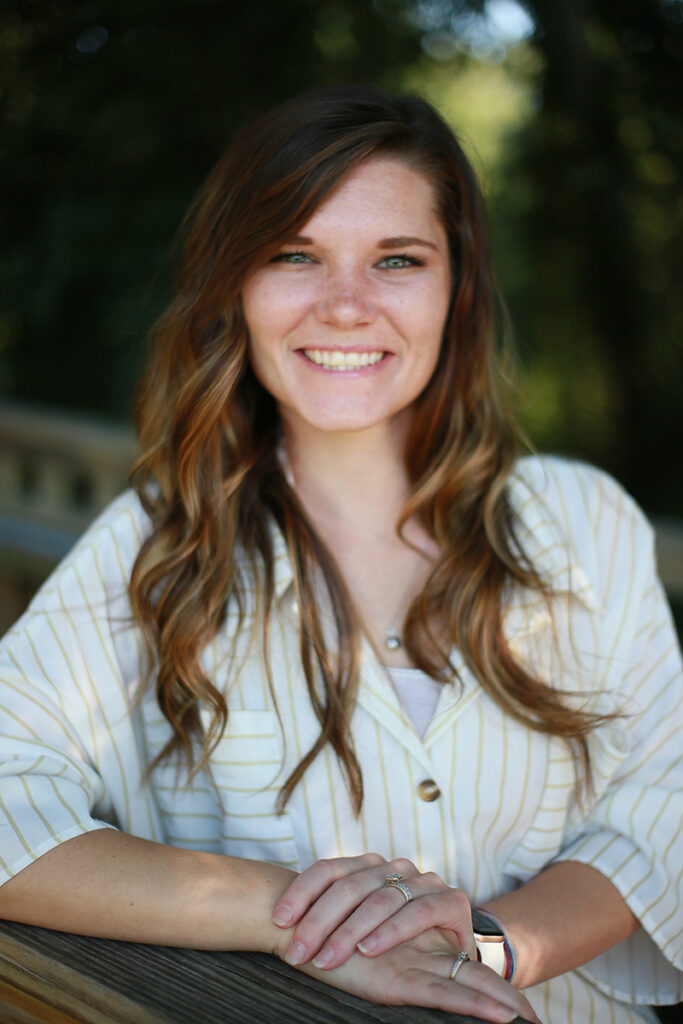 Chelsey O’Neal, a woman with long wavy brown hair wearing a striped shirt, smiles while standing outdoors, resting her arms on a wooden railing.