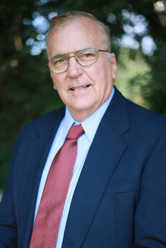 ASAM Certified Jay Weiss, MD, an older man with glasses in a blue suit, light blue shirt, and red tie, stands before a blurred outdoor background filled with greenery.