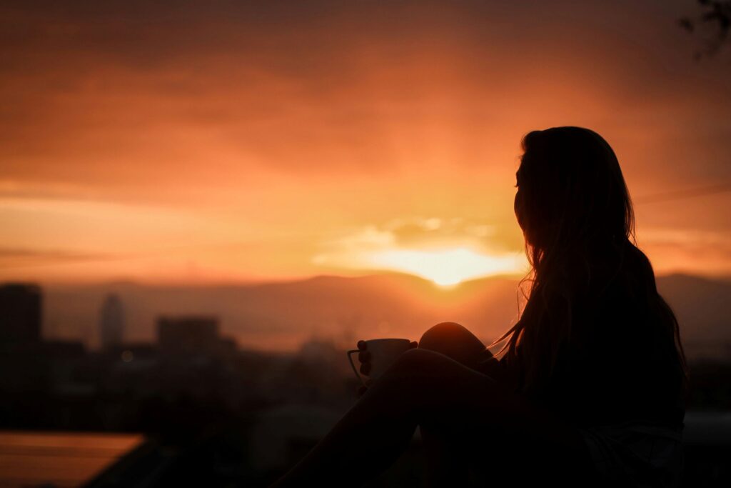 A person in early sobriety sits, holding a cup, gazing toward a vibrant orange and yellow sunset over distant mountains and a city skyline, their silhouette clearly visible in the foreground.