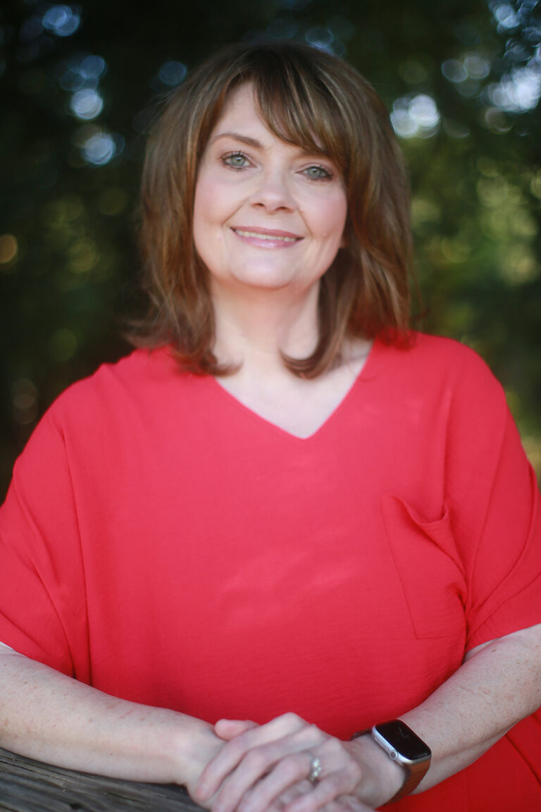 Paulette Grube, RN, with shoulder-length brown hair, stands outdoors in a red blouse, smiling as she rests her hands on a wooden railing.
