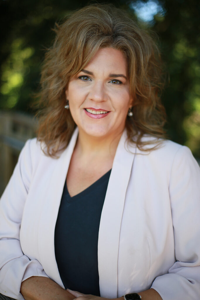 Jennifer Garrett, FNP, with wavy light brown hair, smiles at the camera in a light-colored blazer over a dark top, standing outdoors with greenery in the background.