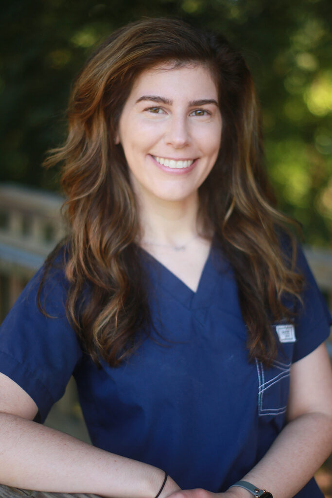 Katie Bedillion, RN, with long brown hair, smiles outdoors in navy blue medical scrubs, framed by trees in the background.