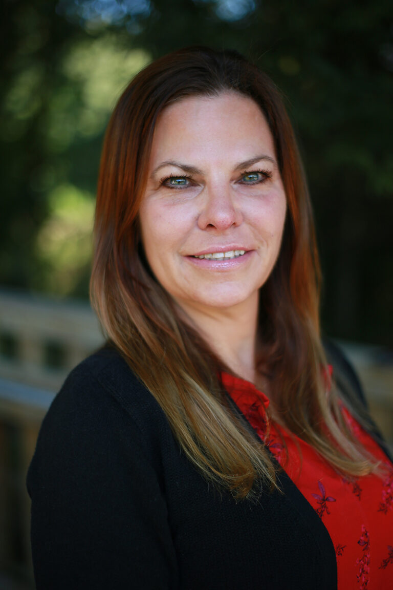 A woman with long brown hair, MA, wearing a red blouse and black sweater is standing outdoors, smiling at the camera. The background is blurred greenery.