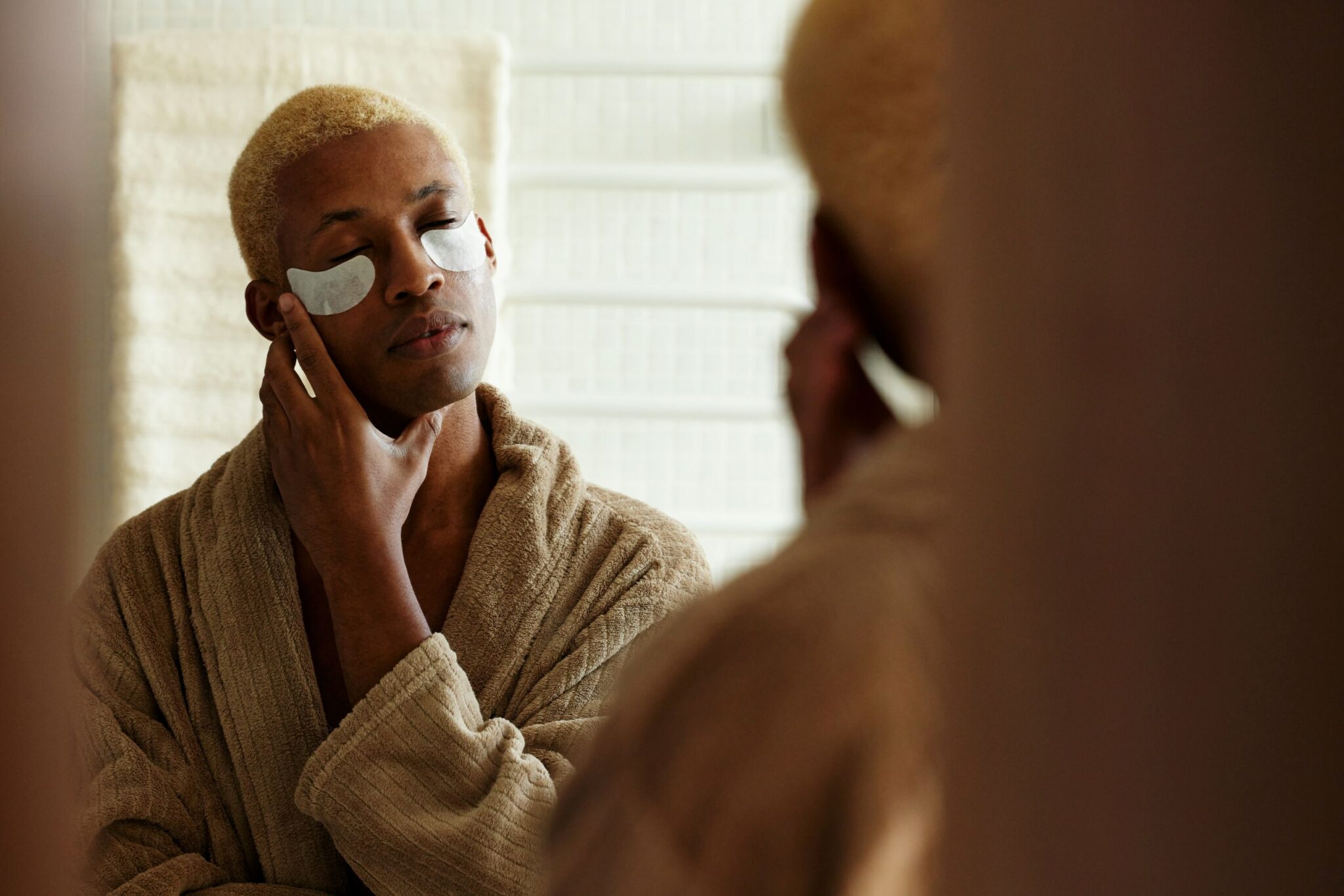 Person with short blonde hair wearing a bathrobe stands in front of a mirror with eyes closed, touching their neck and wearing under-eye patches—embracing a self-care routine as part of their addiction recovery journey.
