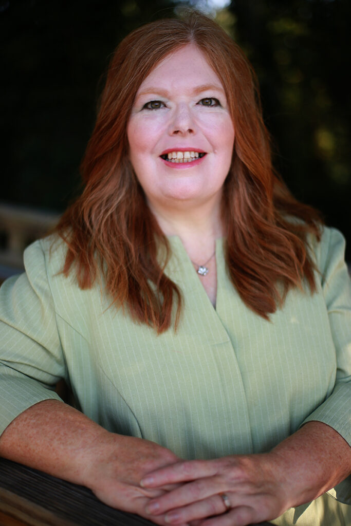 Mary Ann Powell, a woman with long red hair in a light green blouse, smiles as she rests her arms on a wooden surface outdoors, surrounded by greenery.