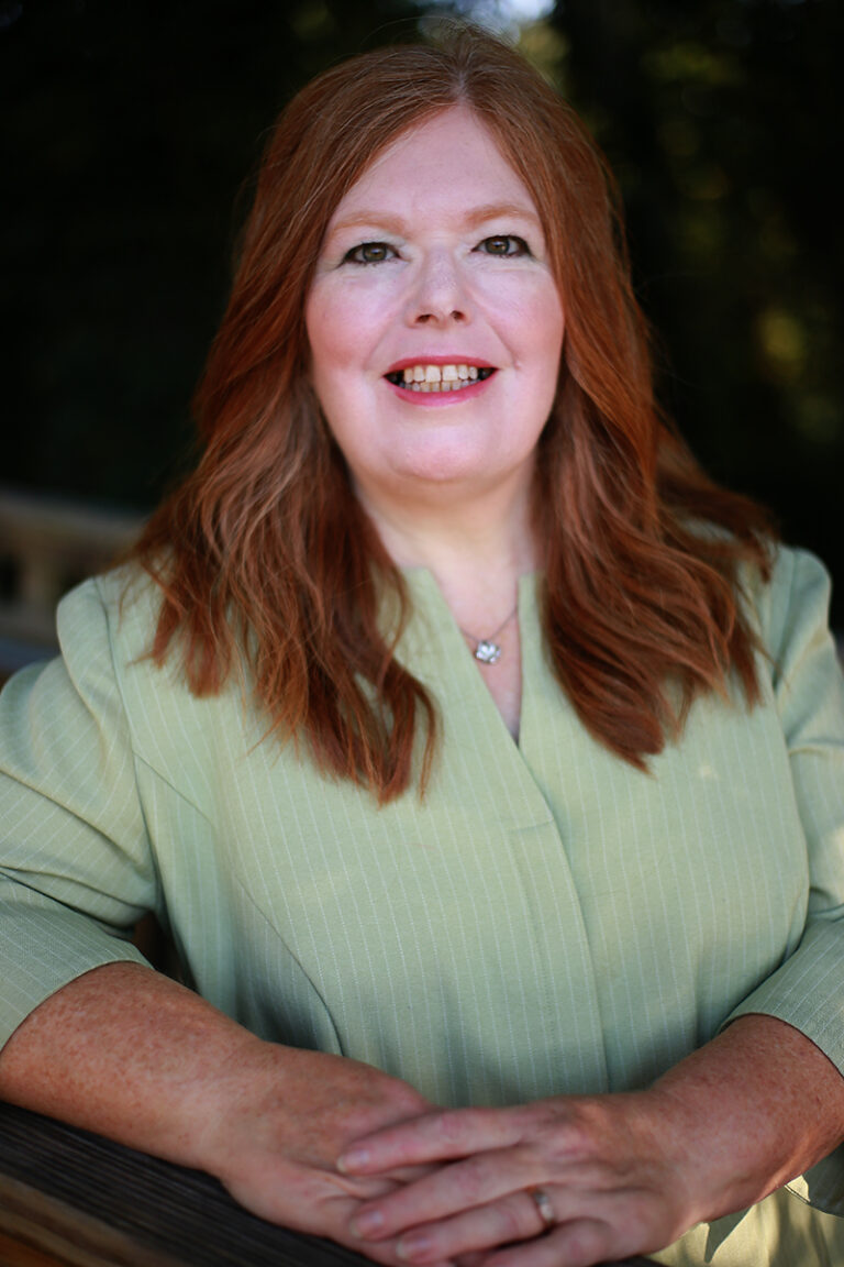 Mary Ann Powell, a woman with long red hair in a light green blouse, smiles as she rests her arms on a wooden surface outdoors, surrounded by greenery.