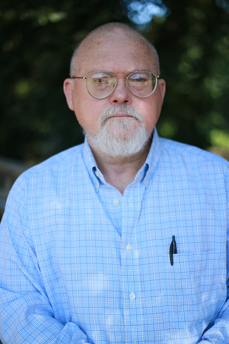 Phillip Ables, LPC, an older man with glasses and a white beard, stands outdoors in a light blue plaid shirt with a pen tucked in the chest pocket.