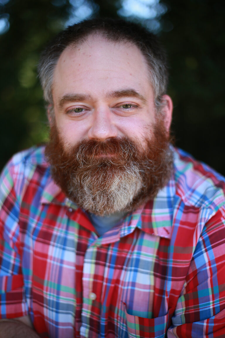 Sean Bryan, LPC, a bearded man with graying hair wearing a colorful plaid shirt, looks at the camera with a slight smile, outdoors with greenery in the background.