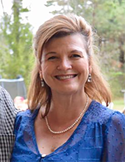 Shirley Ryland, LCSW, with light brown hair wearing a blue blouse, pearl necklace, and earrings, smiles outdoors with trees in the background.