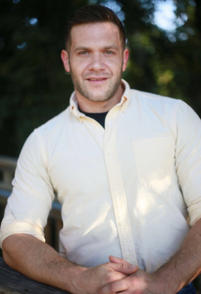 A man with short hair and a trimmed beard, resembling Stuart Bostwick, stands outdoors in a light-colored button-up shirt, resting his hands on a wooden surface—an image capturing the essence of RAC style.