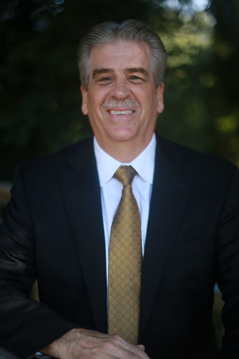 A middle-aged man in a dark suit, white shirt, and patterned tie smiles while standing outdoors with trees in the background, proudly displaying his credentials as an LPC.