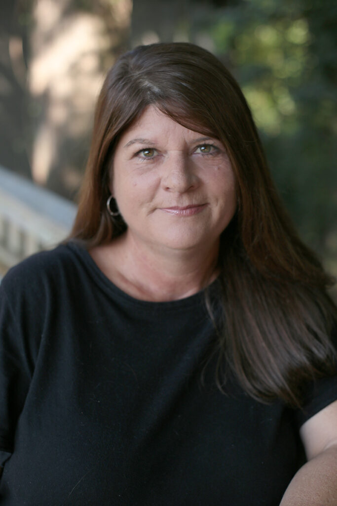 A woman with long brown hair and a black top sits outdoors, looking at the camera with a neutral expression. Trees and a railing are blurred in the background.