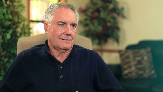 An older man with gray hair sits indoors on a beige chair, wearing a dark blue shirt, with plants and a couch visible in the background.