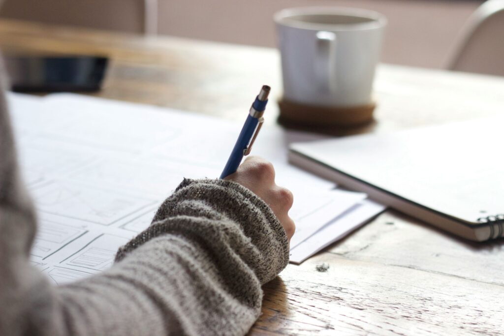 A person wearing a gray sweater writes on papers at a wooden table with a pen. Nearby are a white coffee mug, a closed laptop, and a smartphone—reminders of daily life affected by concerns such as meth abuse.